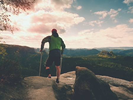 Man with forearm crutches. Hiker after hard walk achieved mountain peak. Tourist with broken leg in immobilizer hold medicine  poles.の写真素材