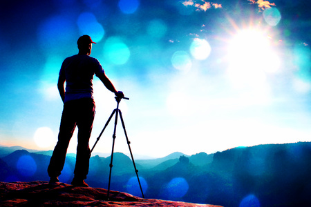 Film grain effect. Professional photographer stay  with tripod on cliff and thinking. Dreamy foggy landscape, blue misty sunrise in a beautiful valley belowの写真素材