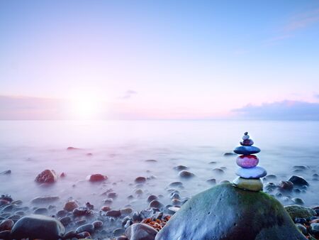 Balanced stone pyramide on shore of blue water of ocean. Blue sky in water level mirror.  Poor lighting conditions.の写真素材