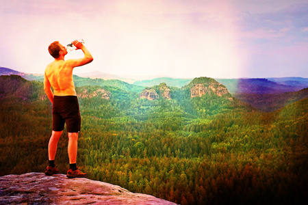 Film grain effect. Thirsty hiker in black pants with bottle of water. Sweaty tired tourist on the peak of sandstone rocky park Saxony Switzerland watching into misty landscape.の写真素材