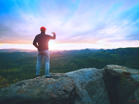 Amateur takes photos with smart phone on peak of rock. Dreamy fogy landscape, spring sunrise in a beautiful valley below, Sun hang on horizonの写真素材