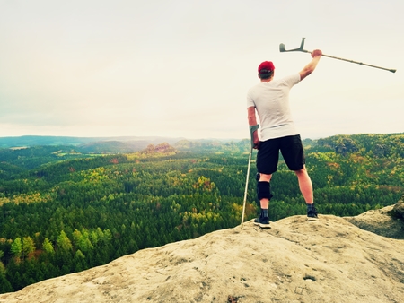 Happy man hiking holding medicine crutch above head, injured knee fixed in knee brace feature. Scenic mountain top with deep cloudy valley belowの写真素材