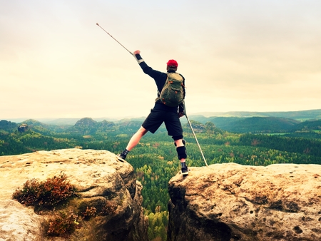 Hiker with broken leg in immobilizer. Tourist with medicine crutch above head achieved mountain peak. Deep misty valley bellow silhouette of happy man with hand in air.の写真素材