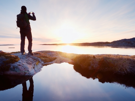 Rear silhouette of travelling man taking selfie at sea. Tourist with backpack standing on a rock, looking  over blue sea and taking photo by his smart phone cameraの写真素材