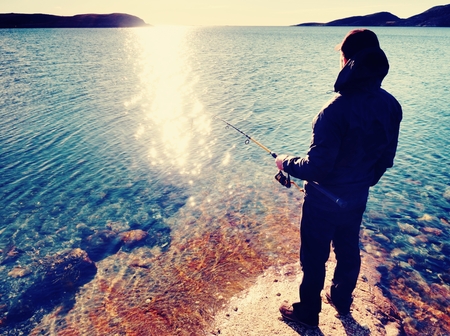 Very close view of working fisherman. Man checking pushing bait and throwing it far into the sea. Fisherman with fishing rod in his hand and stands near water against a beautiful sunset.の写真素材