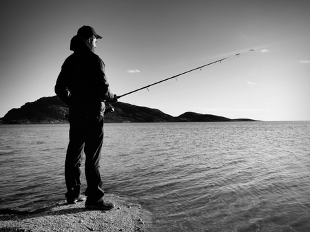 Fisherman check fishing line and pushing bait on the rod, prepare himself and throw lure in peaceful water. The adult man in warm clothes is fishing at the big lake. Fisherman silhouette at sunsetの写真素材