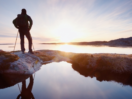 Tall backpacker watch clear sunny spring daybreak over sea. Hiker with backpack stand on rocky shore and his figure is mirrored in water pool.  Hiker enjoy breathtaking sunrise.  Hiking ambition.の写真素材