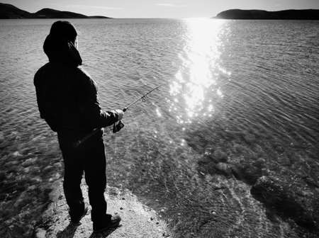The silhouette of a fisherman who throws spinning at dawn. The active man is fishing on the sea from the rocky coast.の写真素材