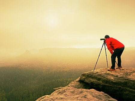 Professional photographer work on mountain peak. Nature photographer takes photos with mirror camera on rock above autumnal landscape. Dreamy fogy  valley below.の写真素材