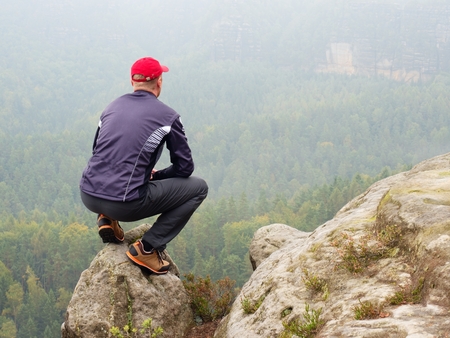 Rear view of alone hiker in dark outdoor suit sit on the edge. Sharp rocky peak  above mountains valley, cold Sun hidden in rainy clouds.の写真素材