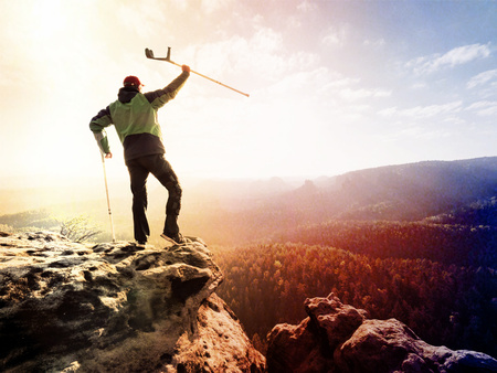 Lens defect. Tourist with medicine crutch above head achieve mountain peak. Hurt backpacker with broken leg in immobilizer stay above valley . The silhouette of man with hand in airの写真素材