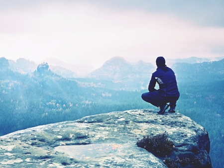 Adult tourist in black trousers, jacket and cap sit on cliff's edge looking to misty hilly valley bellow. Foggy autumn weather. Wet rock withfallen  autumn leavesの写真素材