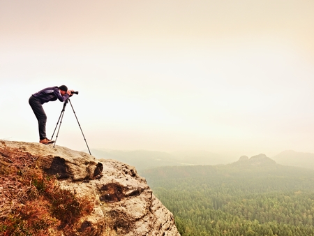 Nature photographer prepare camera to takes impressive photos of misty fall mountains. Tourist photographer at sharp rocky edge on high view point, misty forest bellow.の写真素材