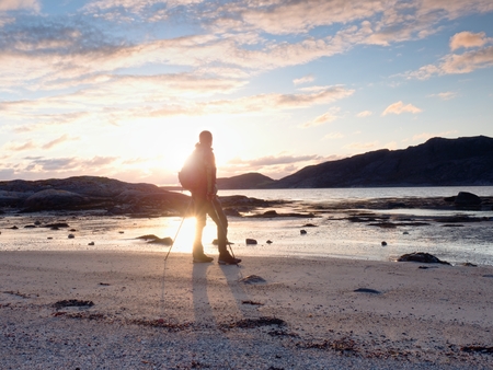 Walker watch sunny spring daybreak over sea. Hiker with backpack stand on sandy shore. Sun rays are  mirrored in water and wet sand. Hiker enjoy breathtaking sunrise.  Hiking ambition.の写真素材