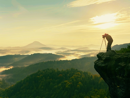 Photographer  on cliff. Nature photographer takes photos with mirror camera on peak of rock. Dreamy fogy landscape, spring orange pink misty sunrise in a beautiful valley below. の写真素材