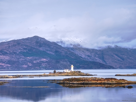 Iasle Ornsay Lighthouse built on a small islet  located on the ferry route. Low level of smooth water. Snowy mountain peaks hidden in cloudy background.  の写真素材