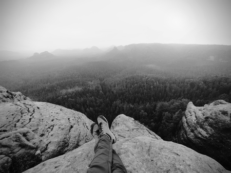 Male legs in dark hiking trousers and leather trekking shoes on peak of rock above  misty valley. Outline of hill side on horizon, Sun hidden in misty clouds.の写真素材