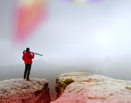 Hiker with camera on tripod takes picture from rocky summit. Alone photographer at edge photograph misty landscape, forest in valley. の写真素材