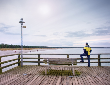 Man thinking. Turtle in warm clothes on sea mole at wooden handrail. Autumn misty day. Tourist on...