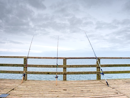 Several fishing rods against the wooden railing of the beach pier. Overcast day, with the hidden sun.の写真素材