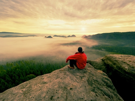 Hiker in black on the rocky peak. Wonderful daybreak in mountains, heavy orange mist in deep valley....