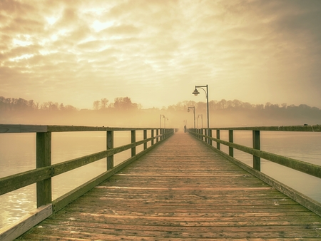 Empty wooden pier in harbor. Steel lamp post, horizon hidden in thick fog. Autumn mist on beach bridge  above seaの写真素材