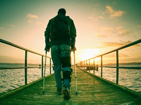 Hurt man with hooded jacket and forearm crutches  standing on sea bridge within  early morning and thinking. Gloomy nostalgic silhouette of sad lonely melancholic adult.の写真素材