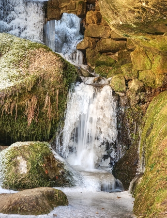 Icicles in frozen waterfall. Snowy and icy stones and boulders with drops of fallen chilly water between sandstone rock.の写真素材