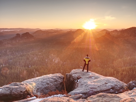 Man on top of mountain. Hiker climbed on peak of rock above foggy valley. Tourist alone looking over landscape at sunsetの写真素材