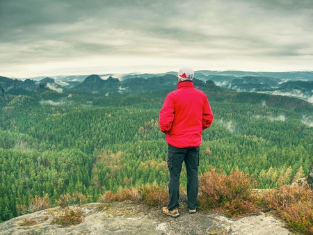 Man tourist on cliff. Hiker in red jacket climbed up to peak enjoy view.  Mountain Climber in warm red jacket  watch the hilly landscapeの写真素材