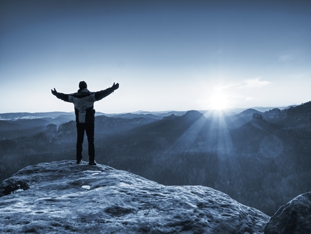 Man on top of mountain. Hiker climbed on peak of rock above foggy valley. Tourist alone looking over landscape at sunsetの写真素材