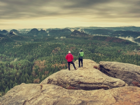 Man hiker in dark sportswear and poles stand on mountain peak rock. Exposed sandstone cliff above forest valley in national parkの写真素材