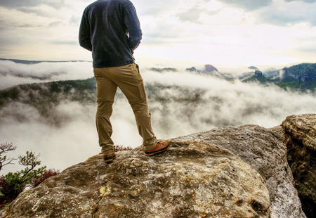 Landscape photograper with camera ready in hand. Man climbed up on exposed rock for fall photos with his digital camera. Cliff above misty autumn landscape.の写真素材
