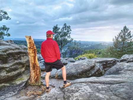 Adult hiker in red shirt and dark running pants. Tall man on the peak of sandstone cliff watching down to landscapeの写真素材