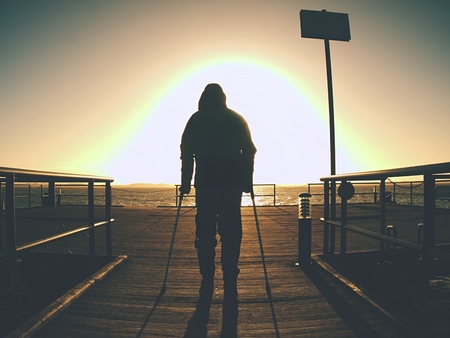 Bended disabled man on coast mole rest on his forearm crutches, warm sunset in backgroundの写真素材