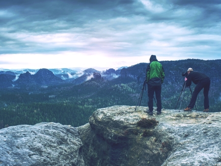 Photographer check display of camera on tripod. Man stay on cliff and takes photos talk friends. Autumn beautiful misty landscape misty sunrise at horizon. Men in mountainsの写真素材