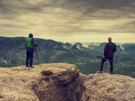 Photographer check display of camera on tripod. Man stay on cliff and takes photos talk friends. Autumn beautiful misty landscape misty sunrise at horizon. Men in mountainsの写真素材