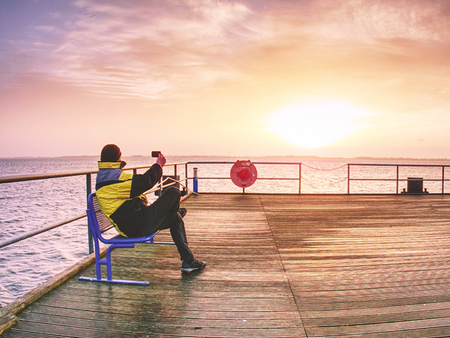 Man photograph morning sea from long wooden mole in wharf. Tourist with smart phone in handの写真素材