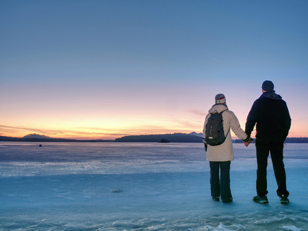 Couple has fun during winter walk on ice of frozen lake. Evening hike at beach of pond. Warm clothes pair with high warm boots.の写真素材