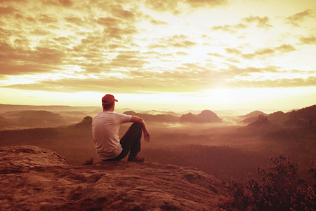 Thinking man sit on rock. Man on exposed summit watching to misty far distance. Cold day in mountains.の写真素材