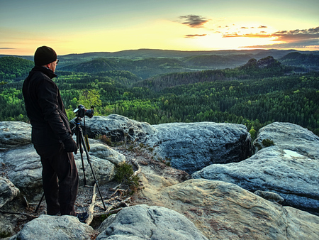 Professional photographer takes photos with camera on tripod on rocky peak Tourist with camera and tripod in cold weatherの写真素材