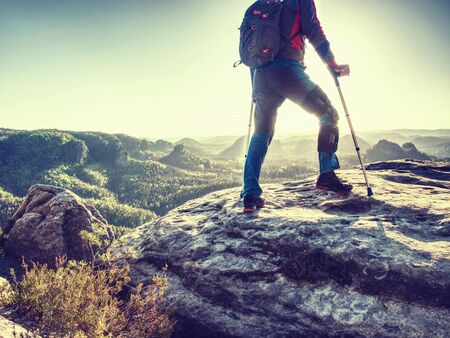 Happy man hiker hold medicine stick,  injured knee fixed in knee brace feature. Scenic mountain top with deep misty valley bellowの写真素材