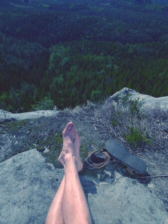 Male hairy legs with sandals rest on rocky summit above valley of natural park. Lazy sunny summer day.