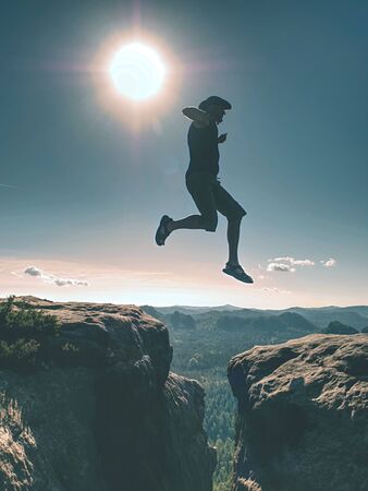 Jumping against Sun. Jumps over cliff. Man on trail through the mountains. Active lifestyle. Man fly over deep gulchの写真素材