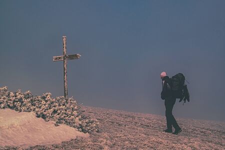 Hiker with backpack reaches the summit of mountain with cross. Snowy freeze mountain peak with meeting place. Success freedom and happiness achievement in mountainsの写真素材