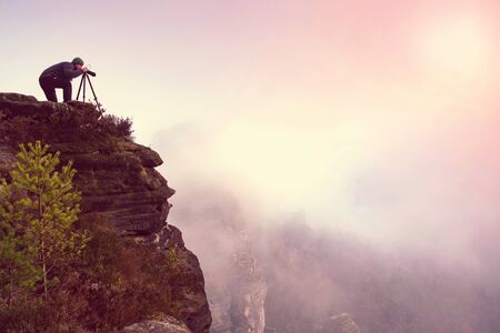 Photographer on mountain cliff looking into mist through viewfinder. Hiker will take picture with camera in hands.の写真素材