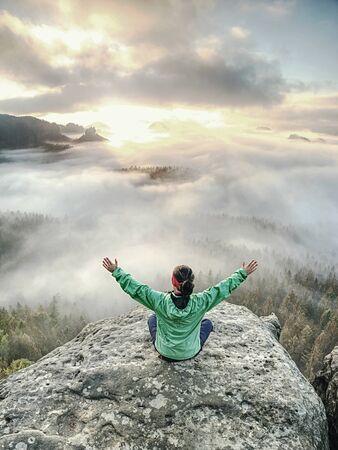 Girl friend sits with raised hands on background of sunrise bay with thick mist and celebrate start...