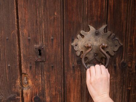 Female hand with black iron knocker on retro old wooden door. The hand knocks on the door. Close up,...
