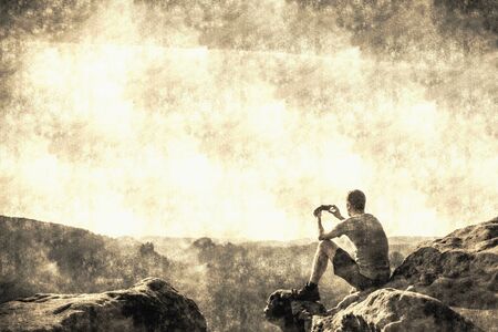 Hiker takes photos by smart phone on peak of mountain. Man in t-shirt and shorts takes photos with smartphone. Dreamy fogy landscape.  Calotype filter. Old photo style.の写真素材