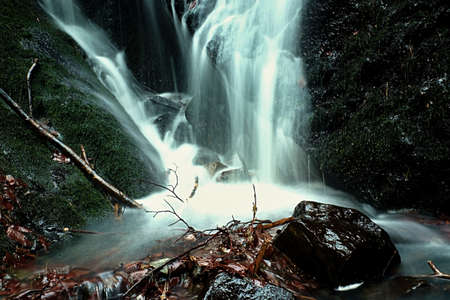 Water spray below small waterfall on mountain stream, water is falling over mossy boulder. The spray...
